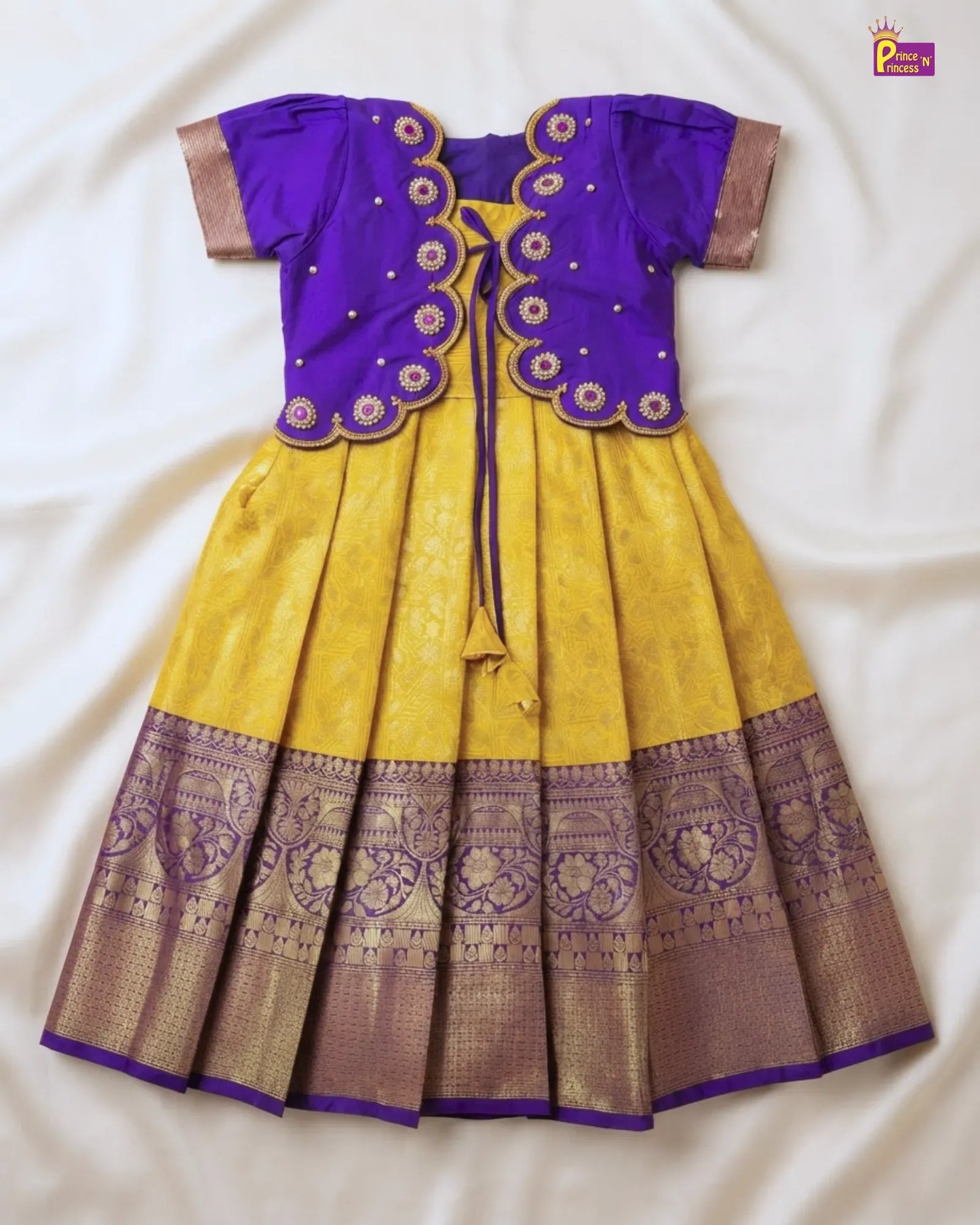 A traditional Indian girl's dress (salwar kameez) featuring a bright yellow skirt with a wide purple and gold zari border, paired with a purple short-sleeve jacket decorated with gold embroidery and scalloped edges.