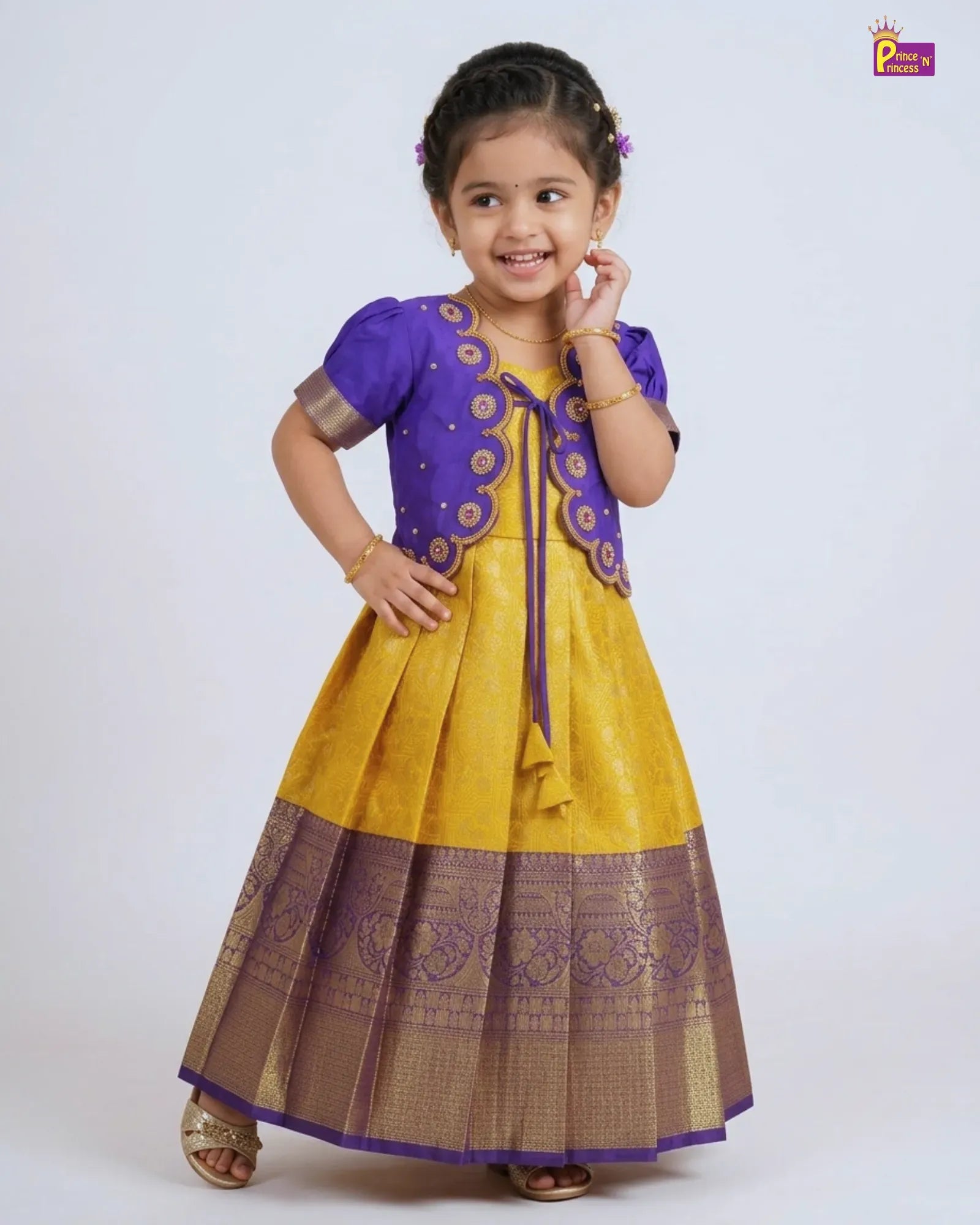 A young girl modeling a traditional Indian outfit consisting of a yellow and purple kurta with a matching pleated skirt and a purple embroidered cardigan. The outfit is set against a plain light gray studio background.