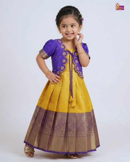 A young girl modeling a traditional Indian outfit consisting of a yellow and purple kurta with a matching pleated skirt and a purple embroidered cardigan. The outfit is set against a plain light gray studio background.