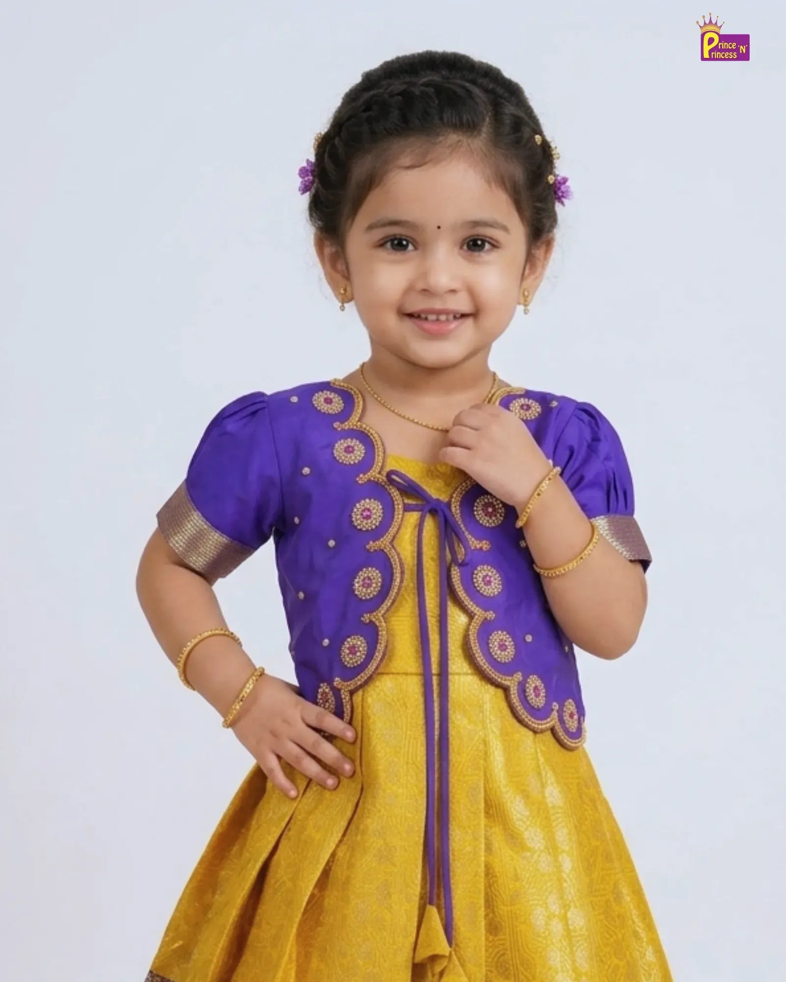 A young girl modeling a traditional Indian outfit consisting of a purple embroidered cardigan over a yellow dress, set against a plain light gray studio background.