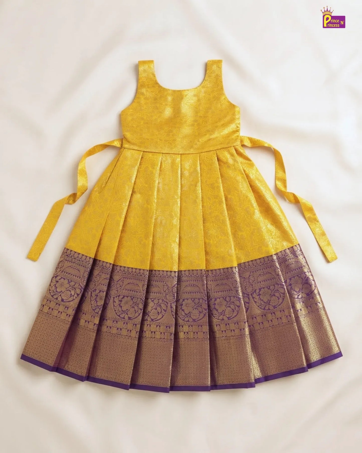 A yellow sleeveless children's dress with a pleated skirt, featuring a wide purple and gold patterned border and matching tie straps, laid flat on a white fabric background.