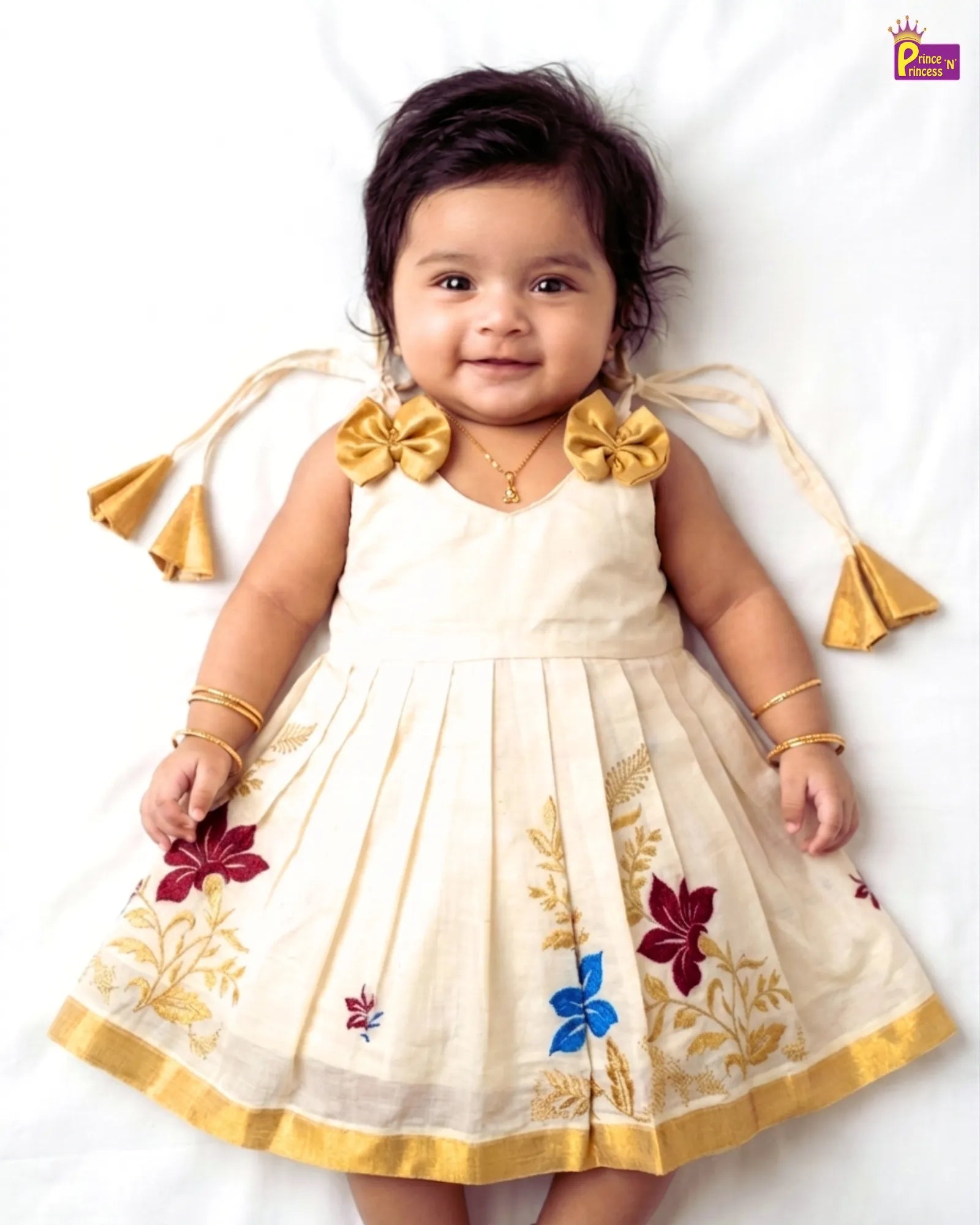 Baby wearing a white dress with floral patterns and gold accents on a white background