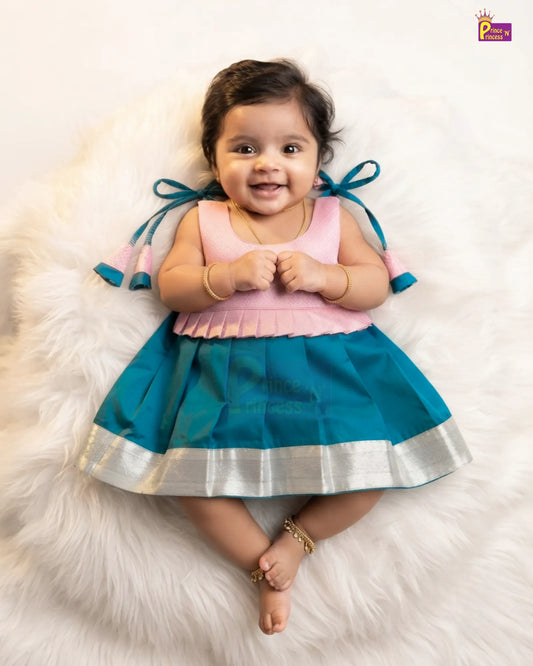 A baby girl wearing a traditional Indian outfit with a pink sleeveless top and a teal blue pleated skirt with a silver border, lying on a white faux fur rug.