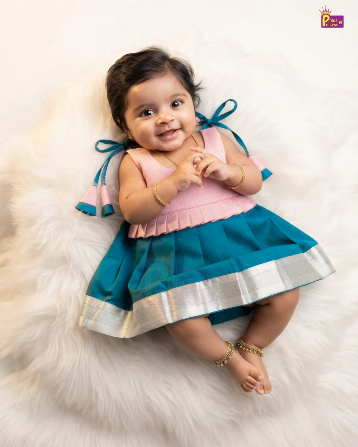 A baby girl lying on a white faux fur rug wearing a two-piece traditional Indian dress with a pink top and teal skirt featuring a silver border.