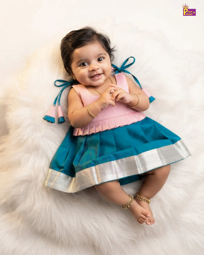 A baby girl lying on a white faux fur rug wearing a two-piece traditional Indian dress with a pink top and teal skirt featuring a silver border.