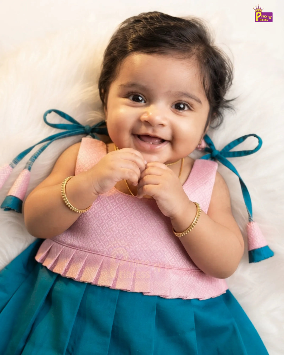 A smiling baby girl lying on a white fluffy surface, wearing a two-tone traditional Indian dress with a pink patterned bodice and a teal blue skirt with bow details.