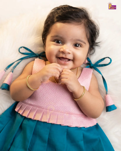 A smiling baby girl lying on a white fluffy surface, wearing a two-tone traditional Indian dress with a pink patterned bodice and a teal blue skirt with bow details.