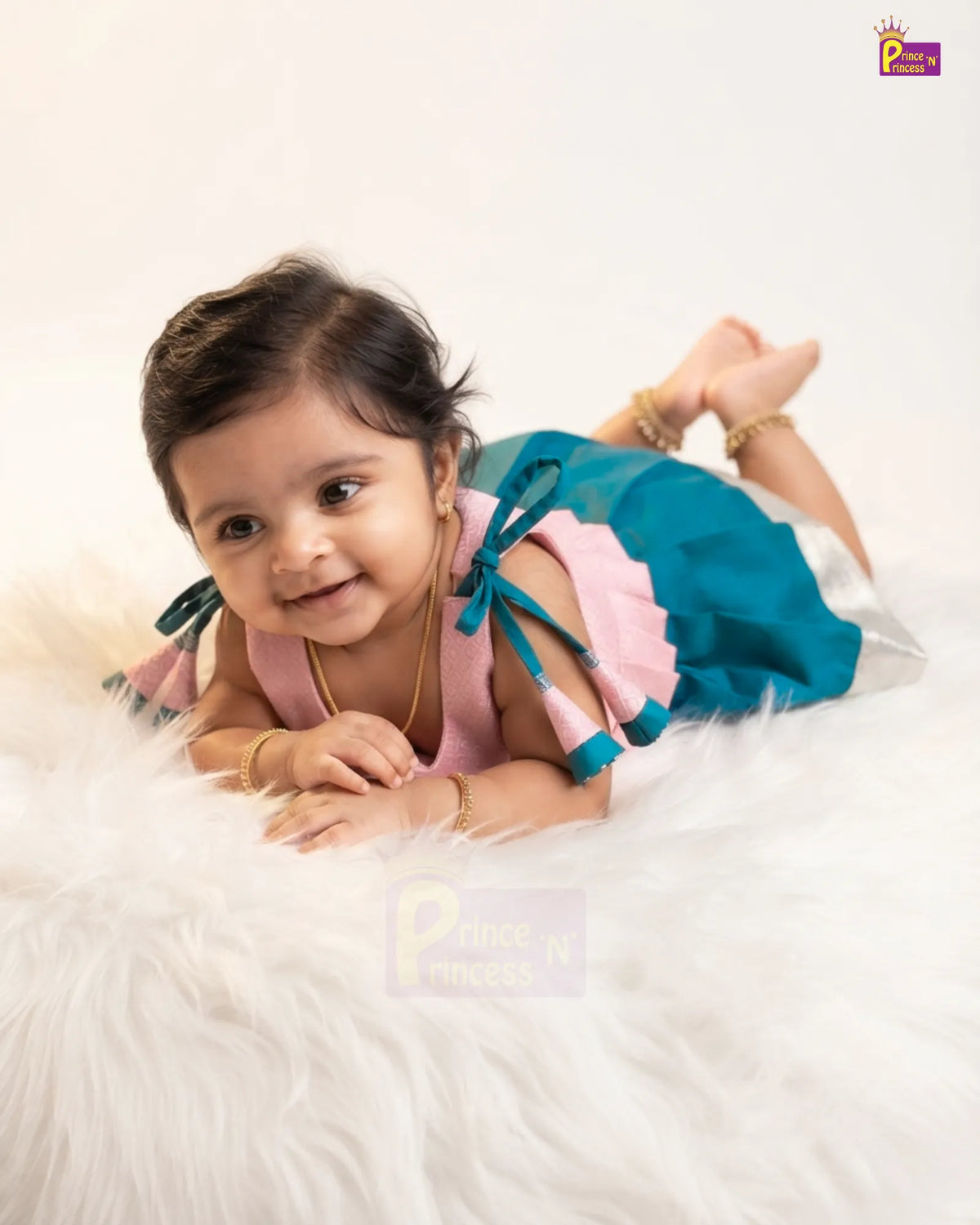 A smiling toddler girl lying on a white faux fur rug wearing a pink and teal dress with gold jewelry, photographed in a studio setting.