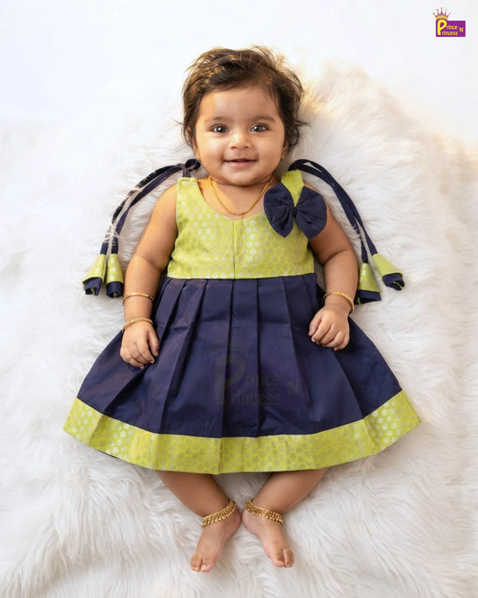 A toddler girl wearing a sleeveless navy blue and lime green dress, lying on a white faux fur rug. The dress features a lime green bodice with a subtle pattern, a navy blue pleated skirt with a matching lime green hem, and a large navy bow on the back of the neck with matching tassels.