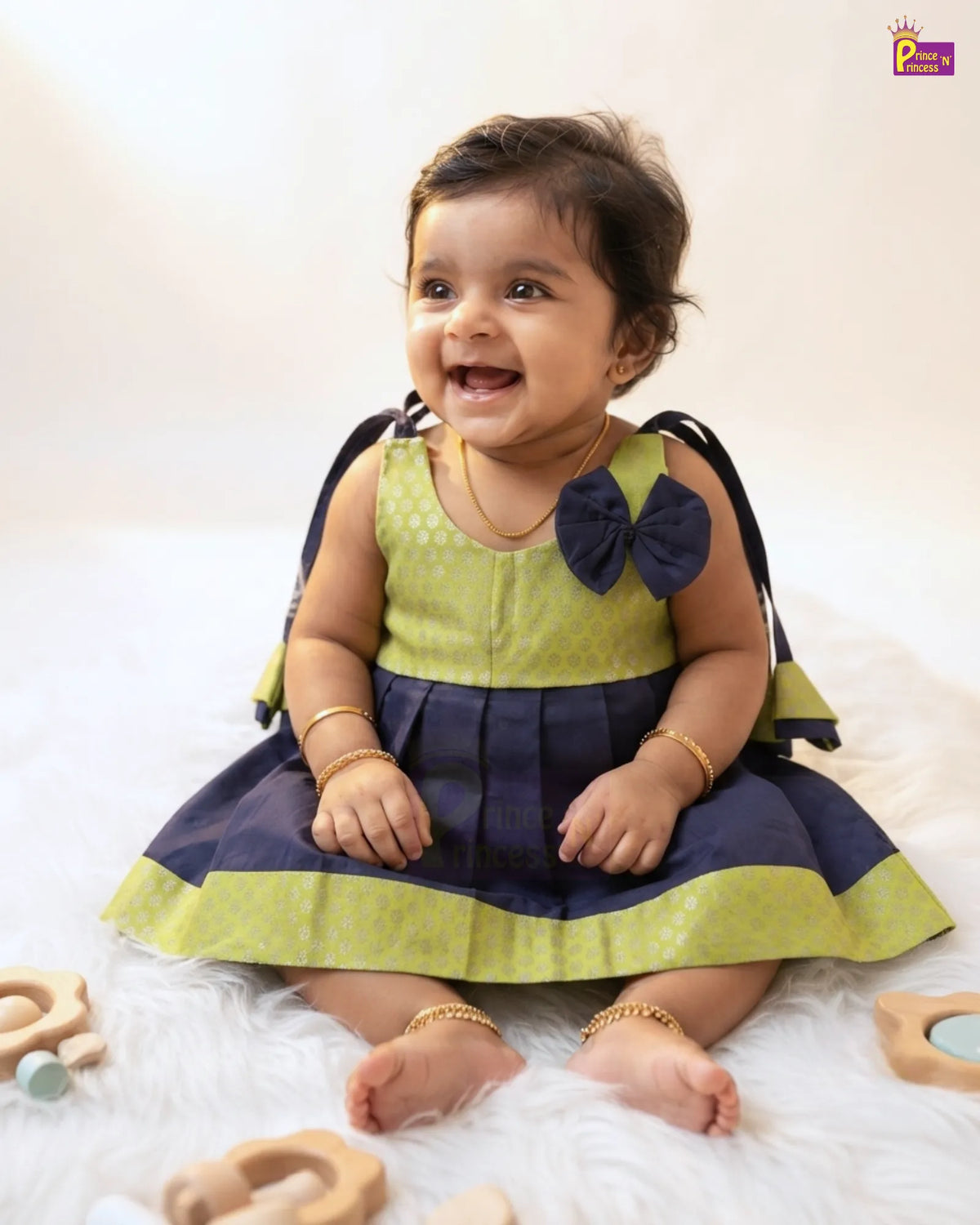 A smiling baby girl wearing a two-tone dress with a lime green top and navy blue skirt, sitting on a white faux fur rug with wooden toys around her.