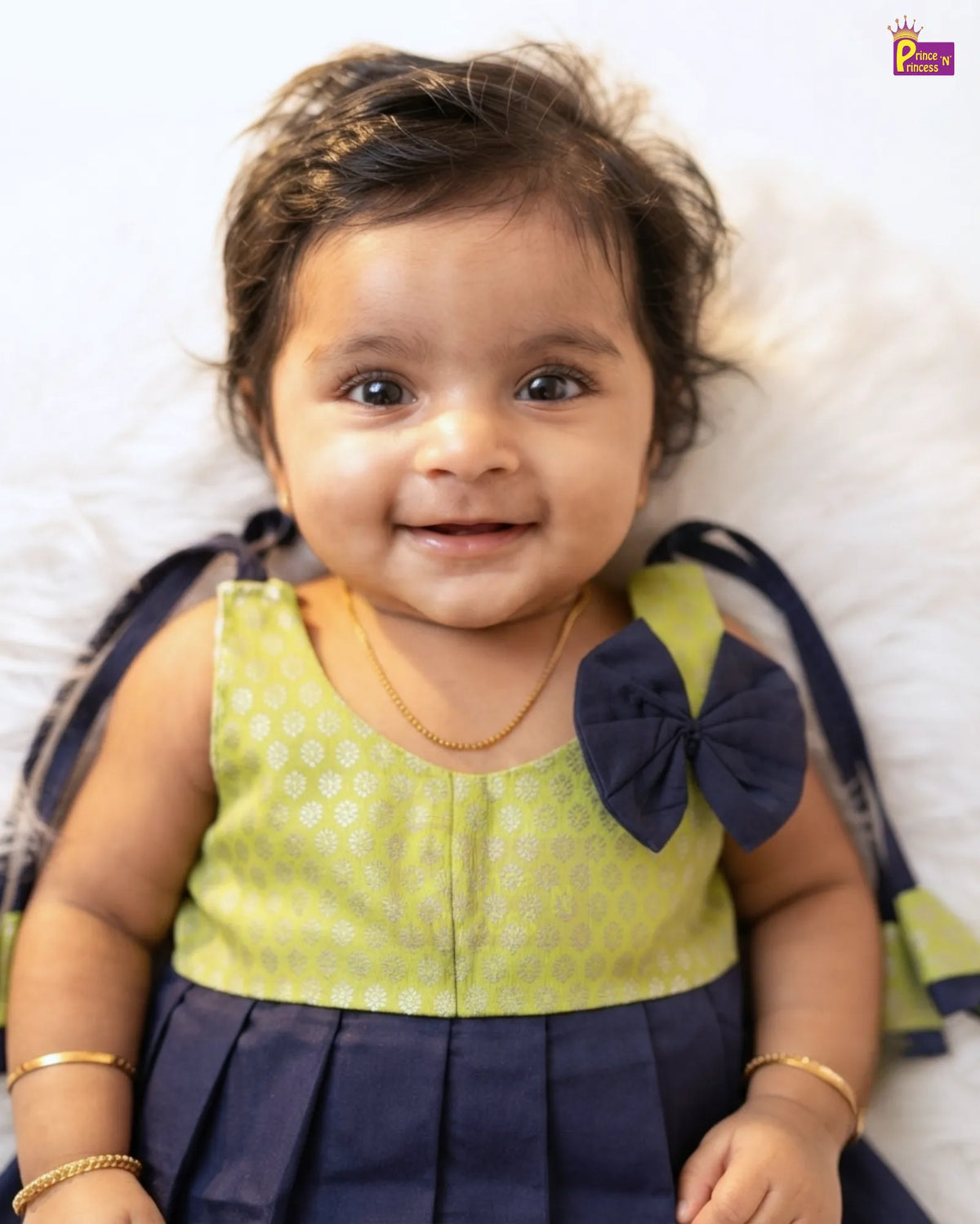 A smiling toddler girl wearing a sleeveless navy and lime green dress with a large bow on the shoulder, lying on a white fluffy surface.