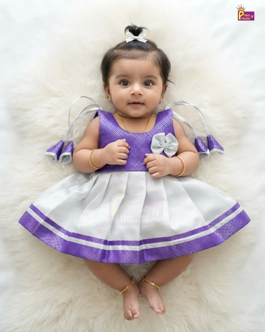 A baby girl wearing a purple and white dress with sheer gray angel wings, lying on a white faux fur rug. The dress features a purple textured bodice, a white pleated skirt with purple and white stripes, and a white bow on the chest.