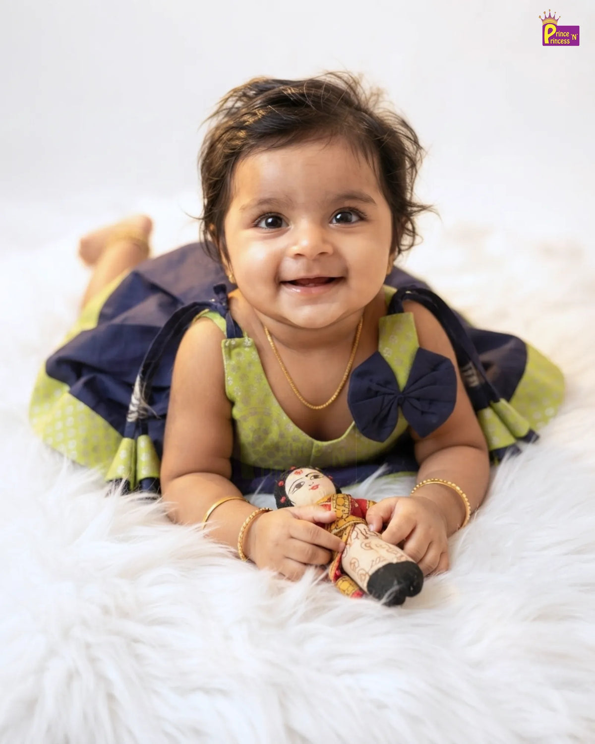 A smiling toddler lying on a white faux fur rug, wearing a green and navy blue dress with a large navy bow on the chest and holding a small doll.