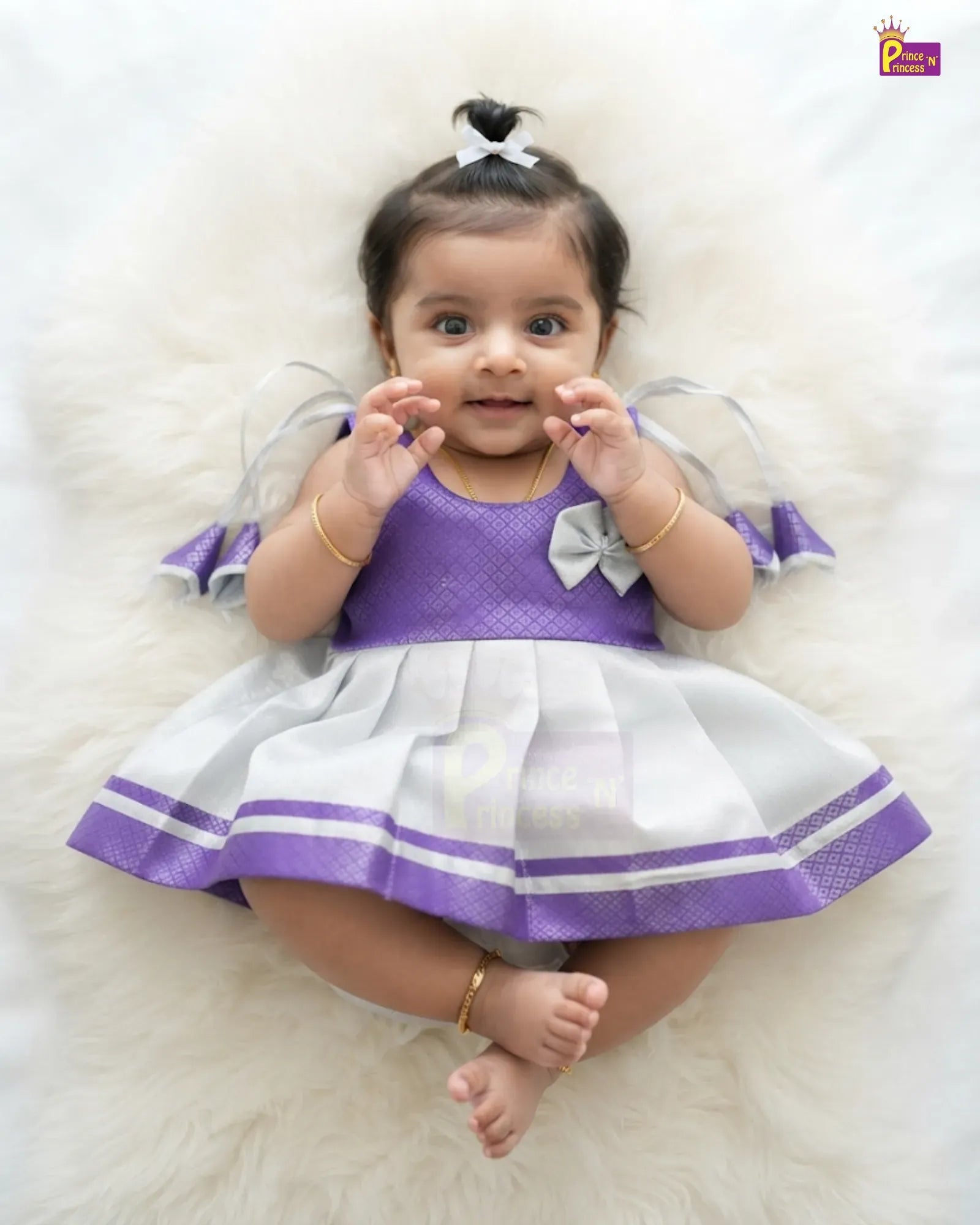 A baby girl wearing a purple and white dress, lying on a white faux fur rug. The dress has a purple textured bodice, white pleated skirt with purple trim, and sheer tulle sleeves with white bows.
