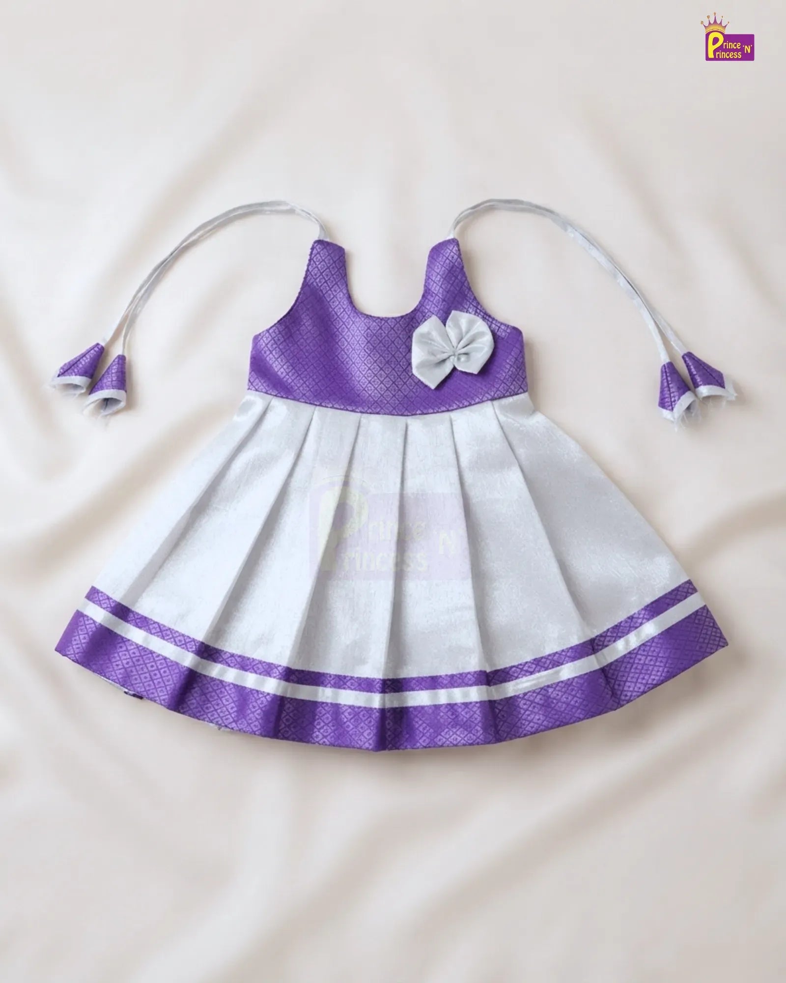 A sleeveless baby girl's dress with a purple textured bodice, white pleated skirt, a white bow on the chest, and matching white and purple tie straps, laid flat on a cream-colored fabric background.