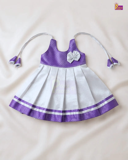 A sleeveless baby girl's dress with a purple textured bodice, white pleated skirt, a white bow on the chest, and matching white and purple tie straps, laid flat on a cream-colored fabric background.