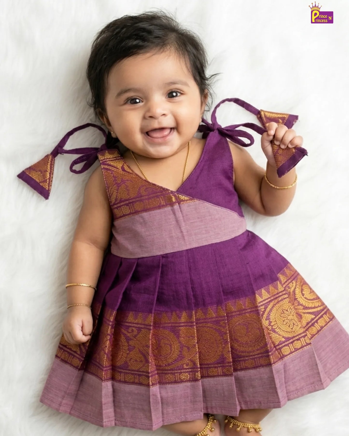 Baby wearing a traditional purple and pink dress with decorative elements on a white background