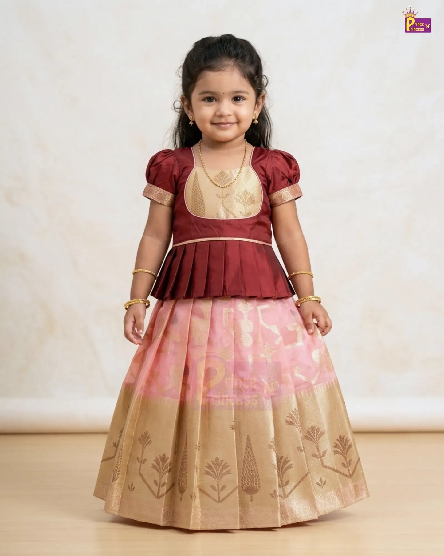 A young girl modeling a traditional Indian ethnic outfit consisting of a maroon and gold peplum top with a pink and gold patterned skirt, set against a neutral beige background.