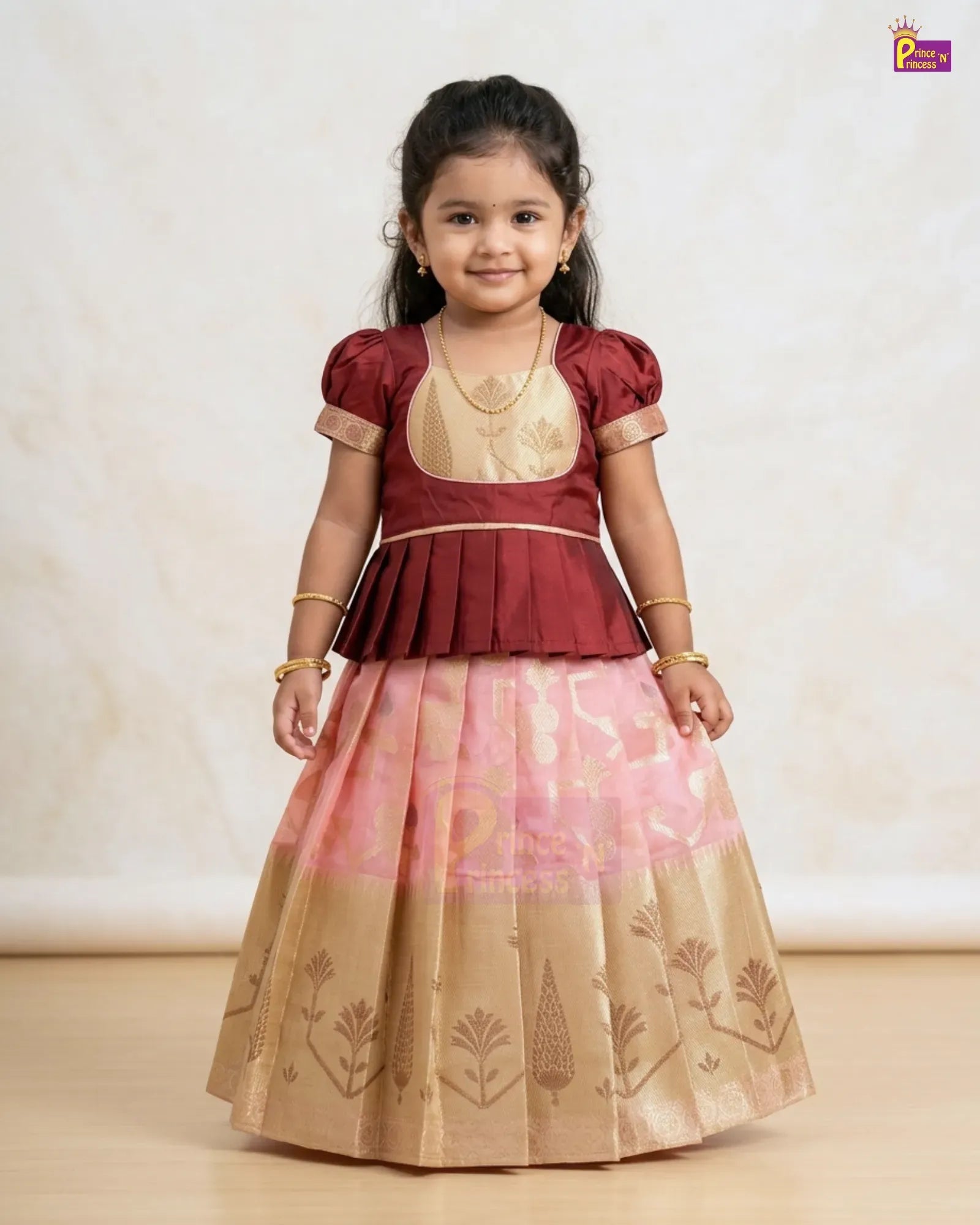 A young girl modeling a traditional Indian ethnic outfit consisting of a maroon and gold peplum top with a pink and gold patterned skirt, set against a neutral beige background.