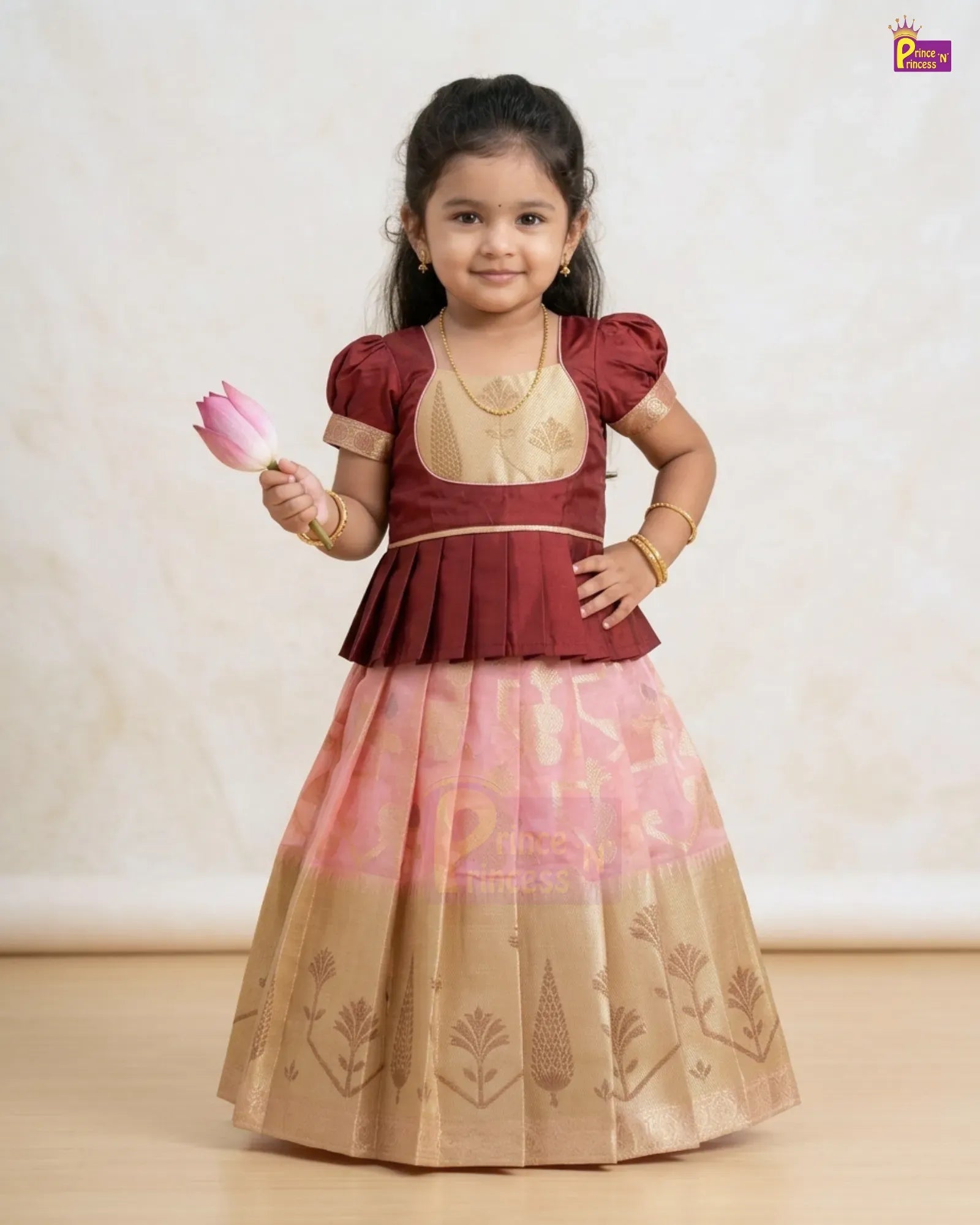 A young girl modeling a traditional Indian ethnic dress, featuring a maroon and beige blouse with a pleated peplum, and a matching pink and beige skirt with gold zari floral patterns. She is holding a pink lotus flower against a neutral beige background.