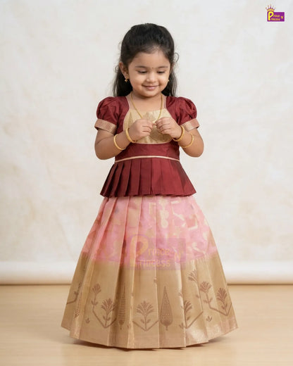 A young girl modeling a traditional Indian ethnic dress with a maroon peplum-style bodice, a light pink upper skirt, and a beige lower skirt with printed motifs, set against a neutral cream background.