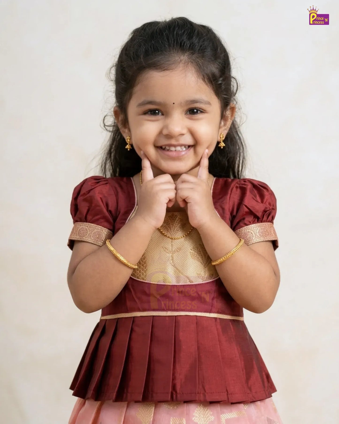 A young girl smiling and posing in a traditional Indian outfit. The outfit features a maroon top with gold embroidery and a pleated skirt in pink and gold.