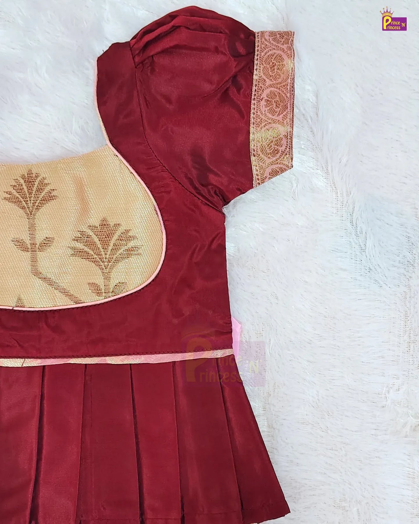A close-up of a traditional Indian outfit, likely a salwar suit or kurta set, featuring a deep maroon red fabric with a pleated skirt, a beige embroidered yoke with gold motifs, and a patterned gold and red border on the sleeve, laid flat on a white textured background.