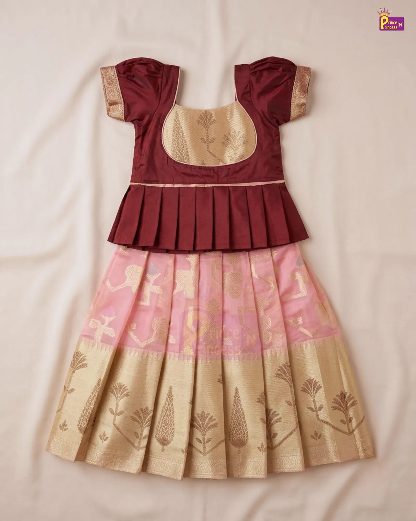 A traditional Indian girl's dress with a maroon peplum top, beige embroidered bodice, and a pleated skirt in pink and beige with gold zari motifs, laid flat on a light beige fabric background.