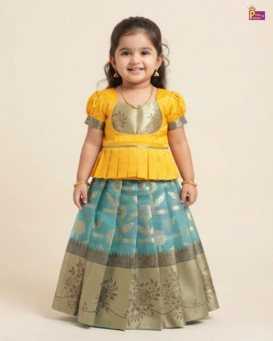 A young girl modeling a traditional Indian ethnic outfit consisting of a bright yellow peplum top with metallic gold embroidery and a matching pleated skirt in turquoise blue and beige with floral patterns.