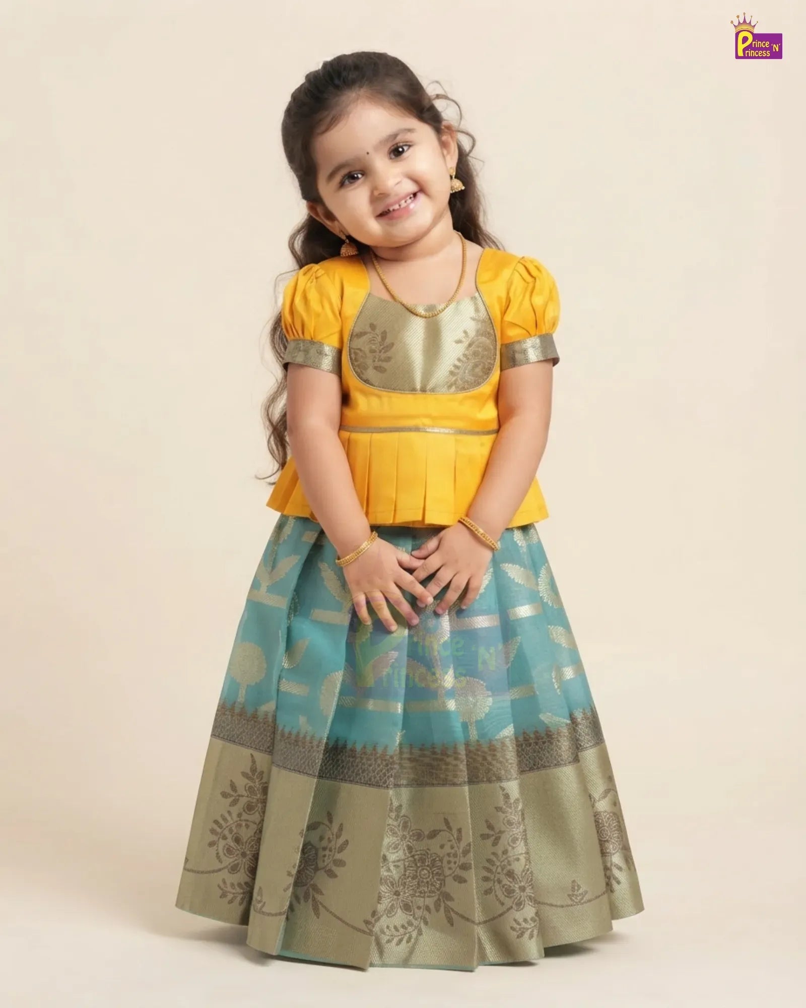 A young girl modeling a traditional Indian outfit consisting of a bright yellow short-sleeve blouse with metallic gold embroidery and a flared teal blue skirt with gold floral patterns and a metallic olive-gold border, set against a plain beige background.