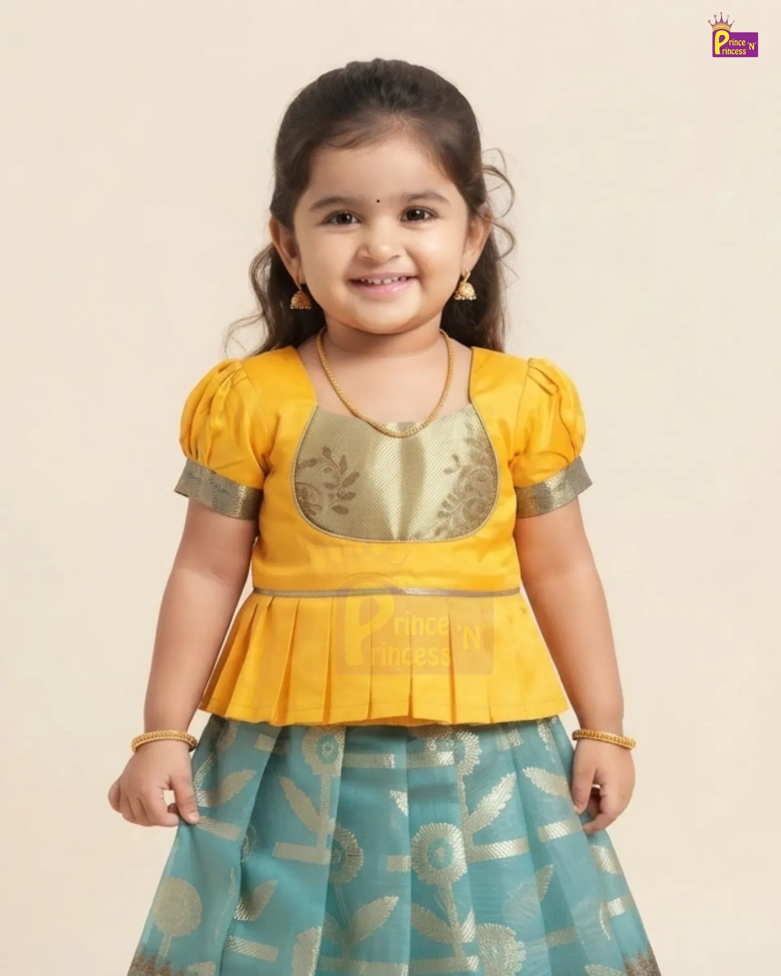 A young girl modeling a traditional Indian outfit consisting of a bright yellow peplum top with gold embroidery and a teal blue skirt with gold patterns, set against a plain beige background.