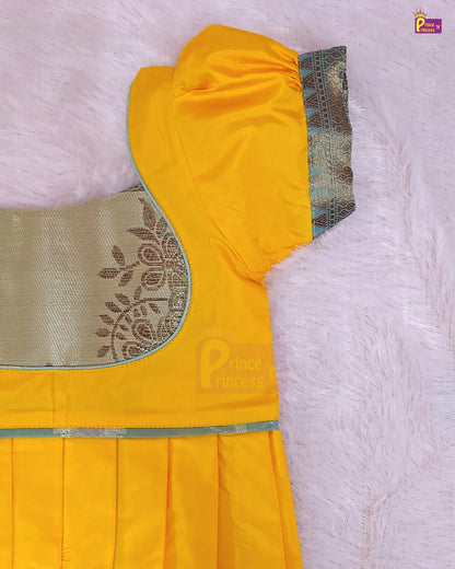 A close-up of a yellow children's dress or kurta laid flat on a soft pink faux-fur surface. The garment has a gold embroidered yoke, a light blue-green border trim, and a pleated skirt.