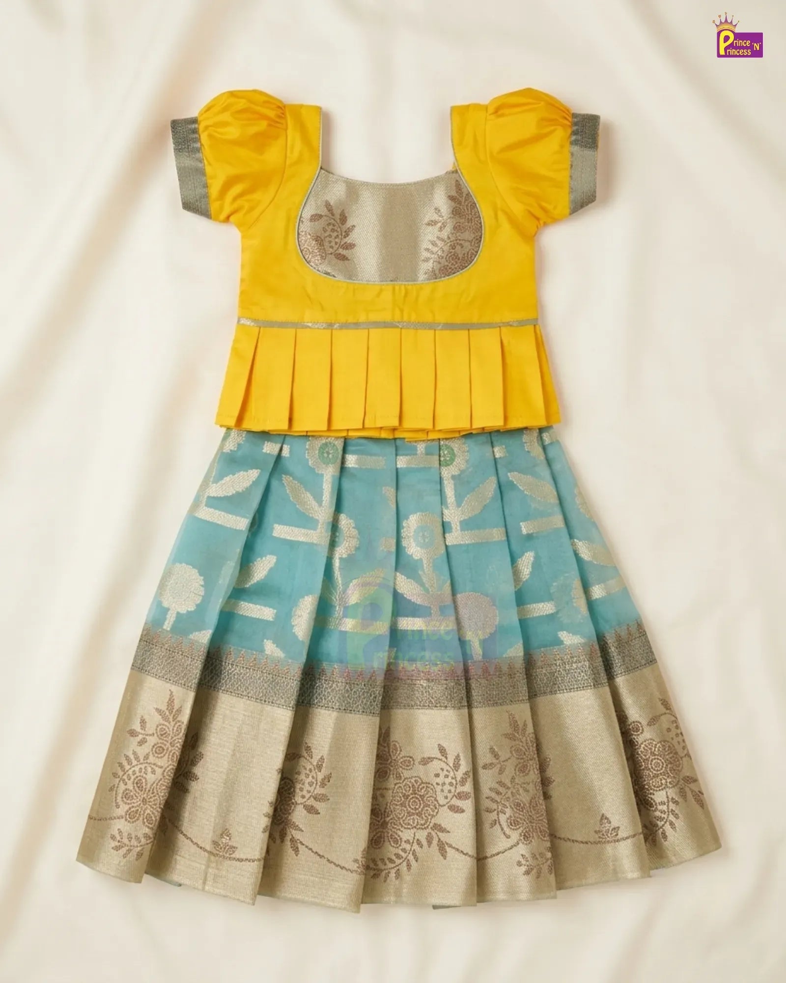 A traditional Indian girl's dress featuring a bright yellow peplum top with grey sleeve cuffs and a light blue pleated skirt with beige floral patterns and a metallic border, laid flat on a cream fabric background.