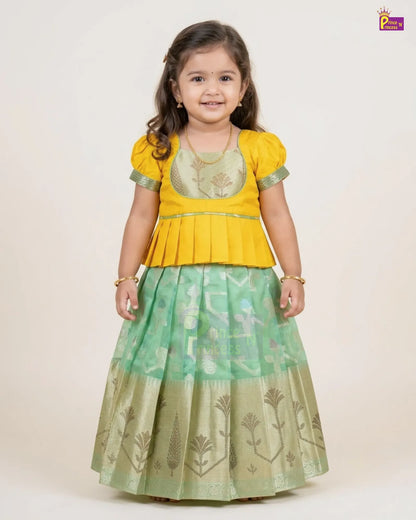 A young girl modeling a traditional Indian outfit consisting of a bright yellow peplum top with green trim and a light green patterned skirt with gold motifs, set against a plain beige background.