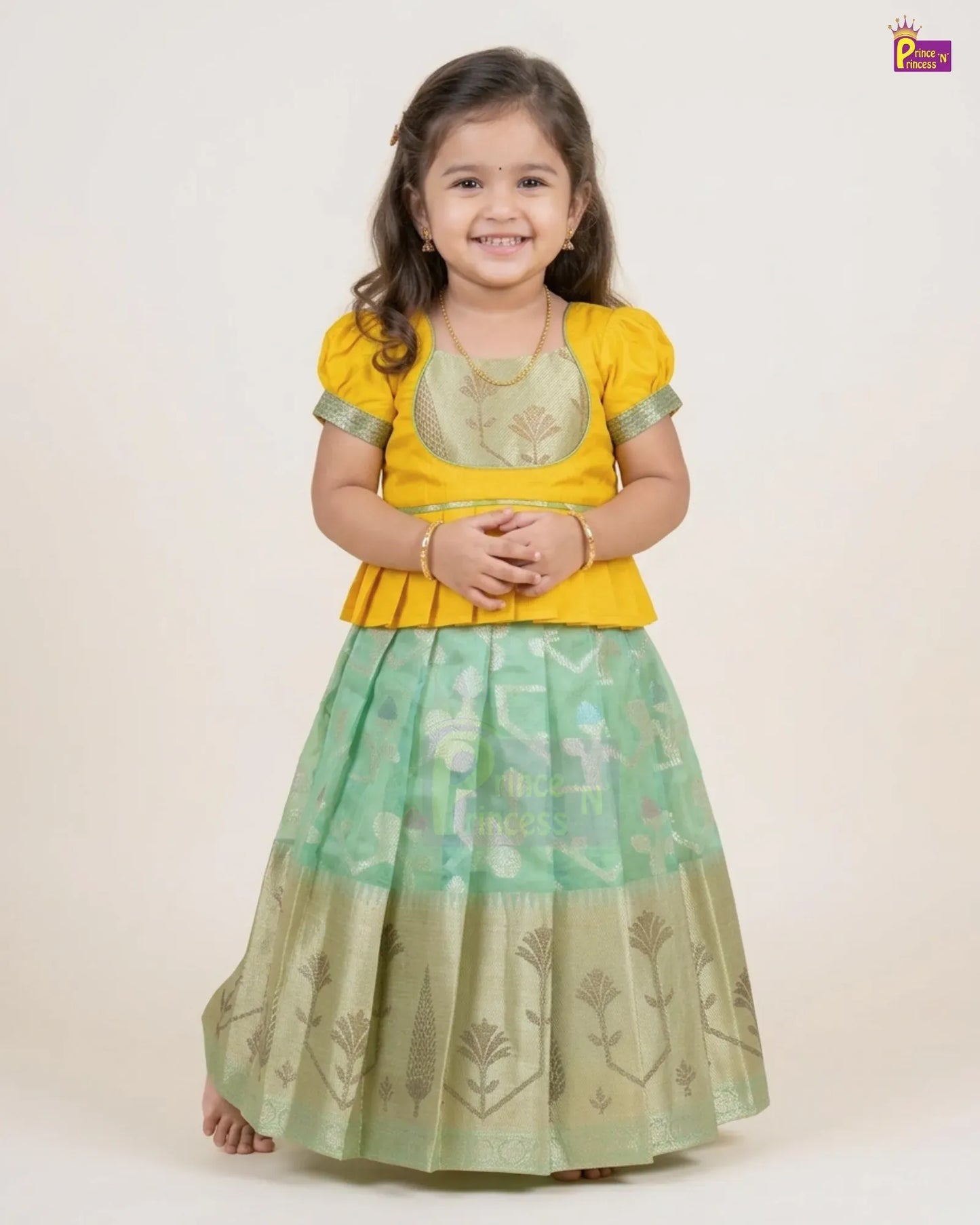A young girl modeling a traditional Indian outfit consisting of a bright yellow short-sleeve blouse with a green embroidered yoke and a pleated light green skirt with gold floral patterns, set against a plain beige background.