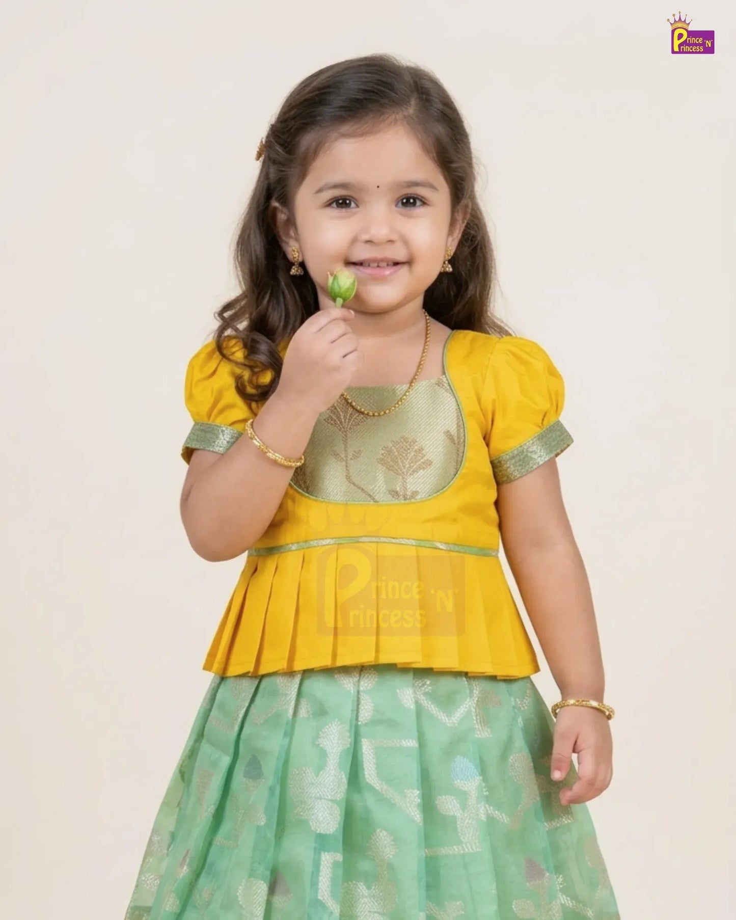 A young girl modeling a traditional Indian outfit consisting of a yellow peplum-style top with green trim and a light green patterned skirt, holding a small green leaf against a plain light beige background.