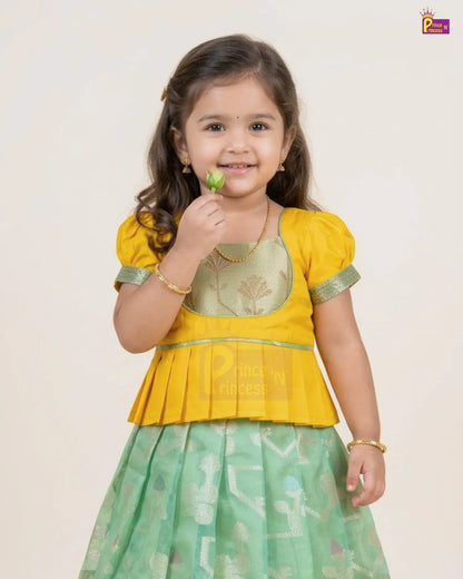 A young girl modeling a traditional Indian outfit consisting of a yellow peplum-style top with green trim and a light green patterned skirt, holding a small green leaf against a plain light beige background.