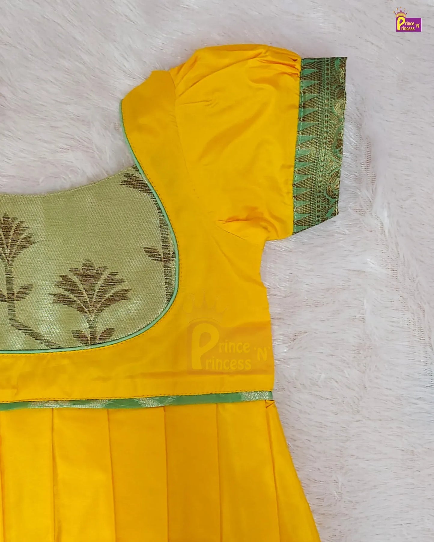 Yellow traditional Indian dress with a green and gold patterned border, a green embroidered yoke, and pleated skirt, laid flat on a white textured background.