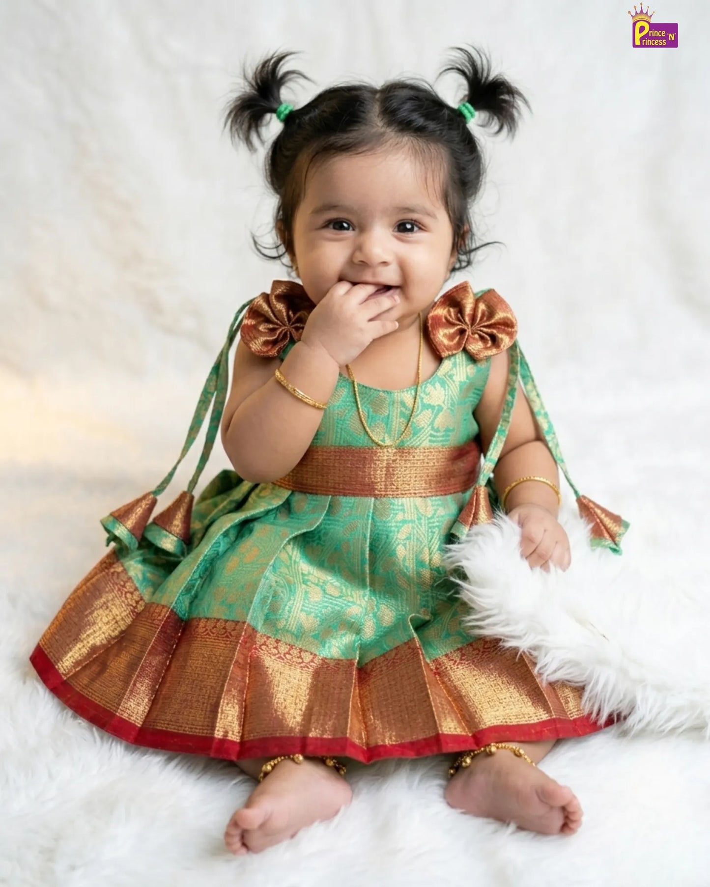 Baby in a green and gold traditional outfit with decorative bows on a white background
