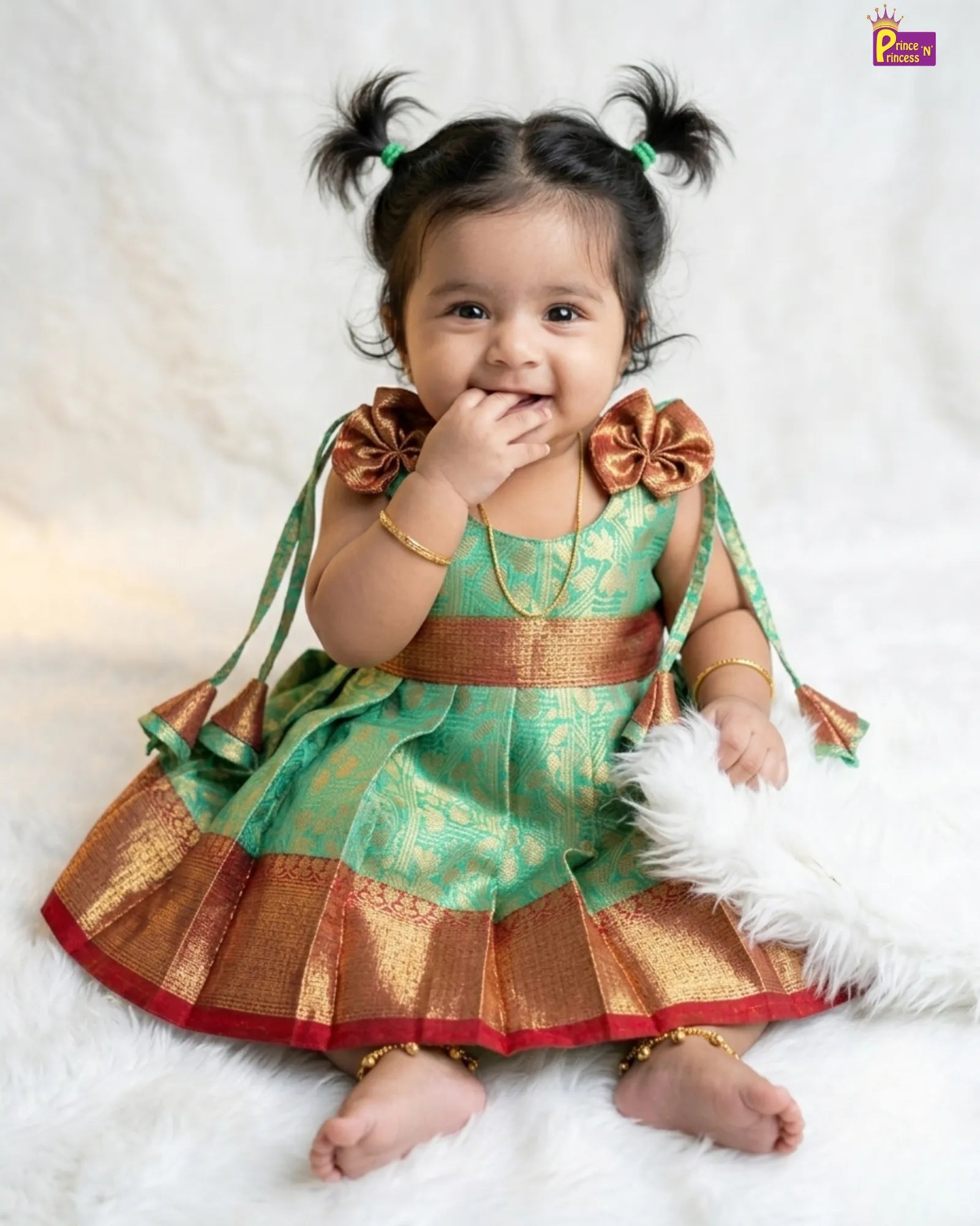 Baby in a green and gold traditional outfit with decorative bows on a white background