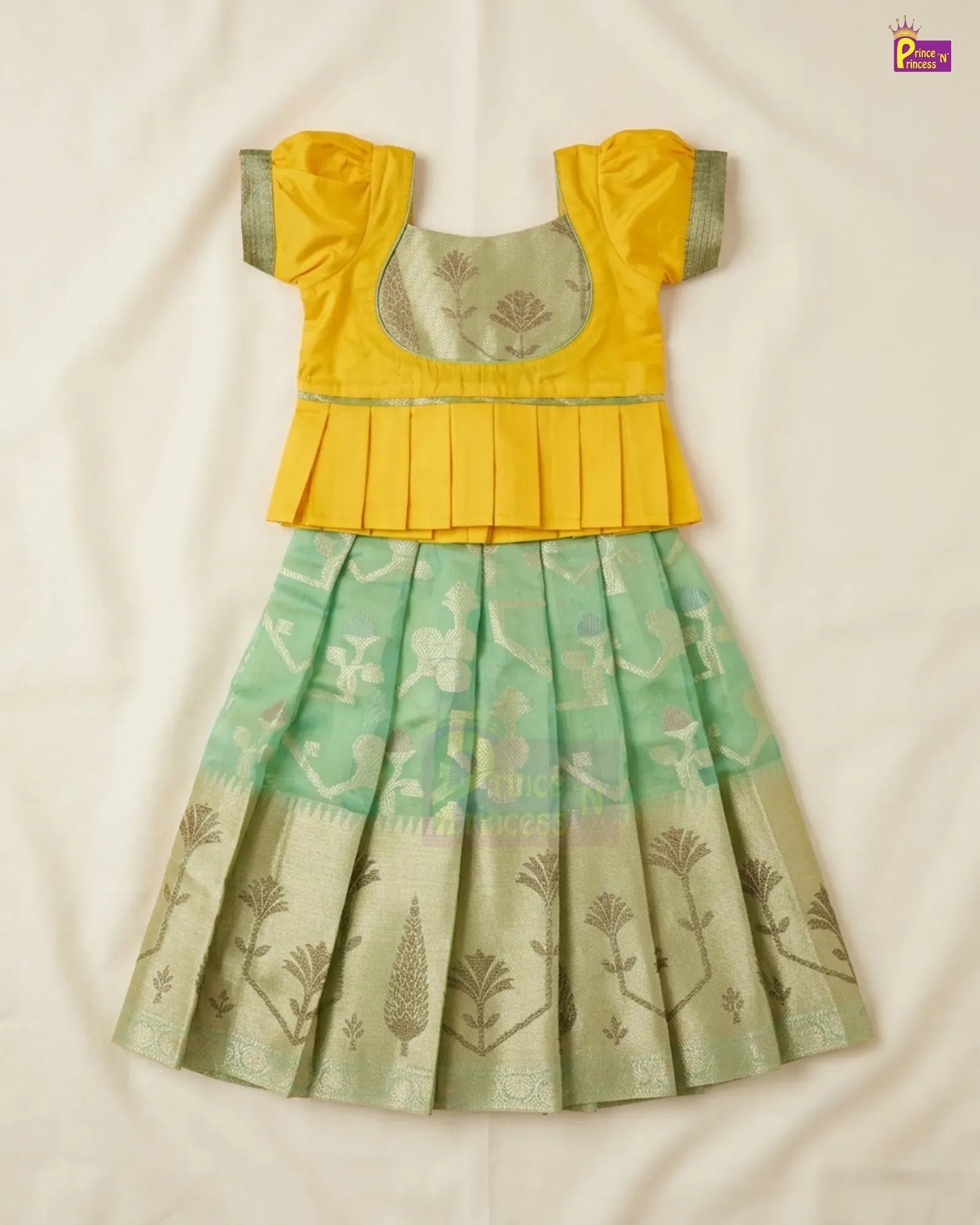 A traditional Indian girl's dress consisting of a bright yellow peplum-style blouse with short puff sleeves and a pleated skirt in mint green and beige with floral motifs, laid flat on a white fabric background.