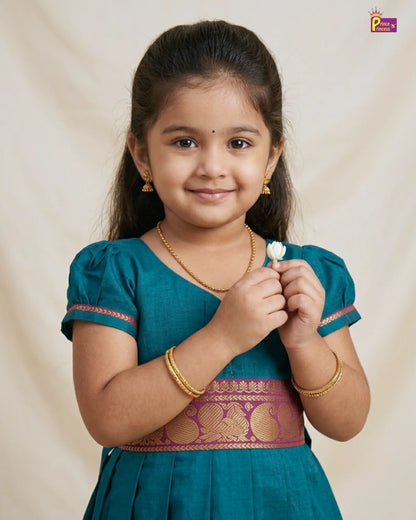 Young girl in a teal traditional outfit holding a white flower against a beige background