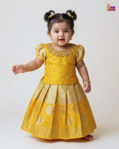 A toddler girl wearing a yellow traditional Indian dress with gold embroidery and a pleated skirt, standing against a plain light background.