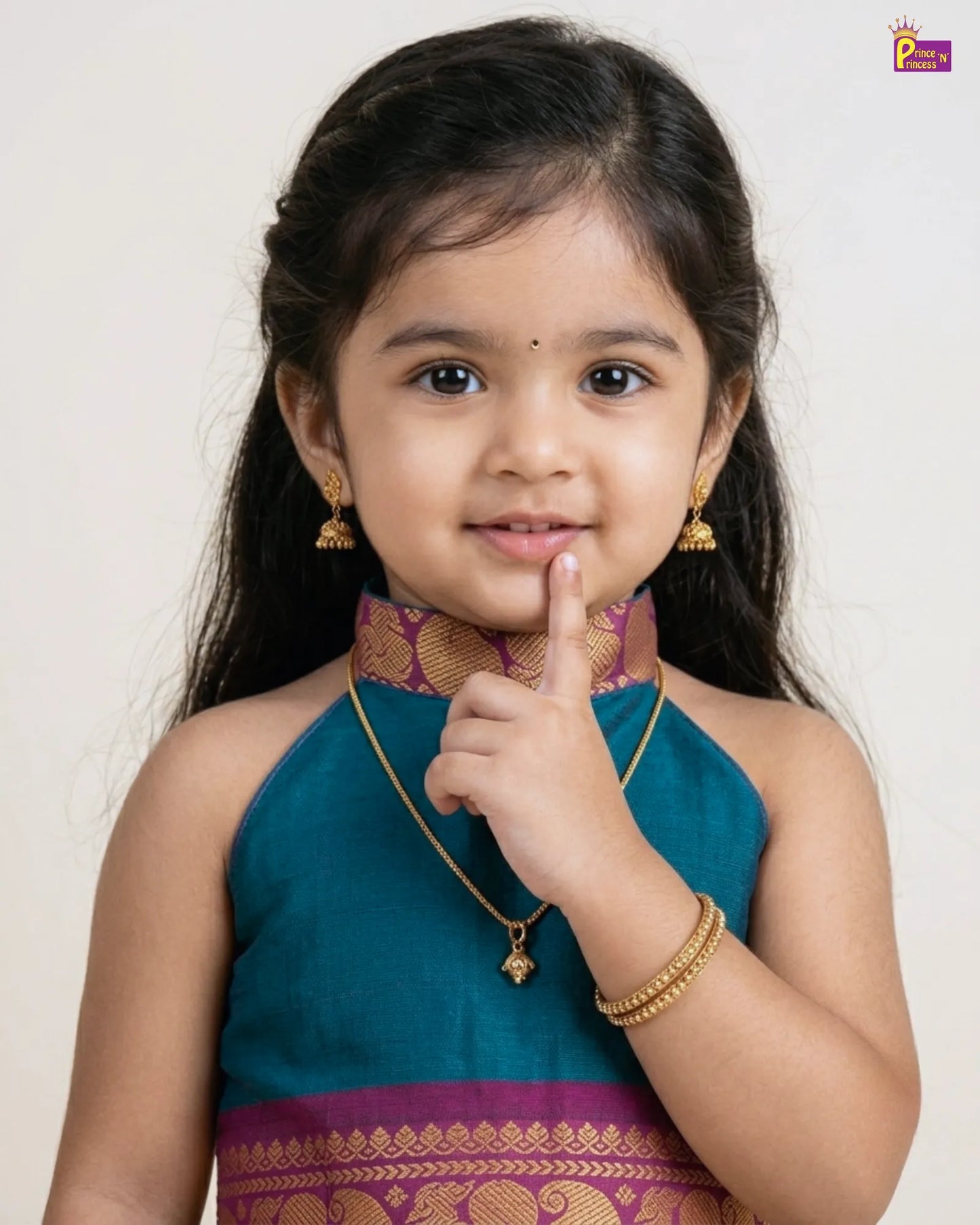Young girl in traditional attire with jewelry, pointing to her chin against a plain background