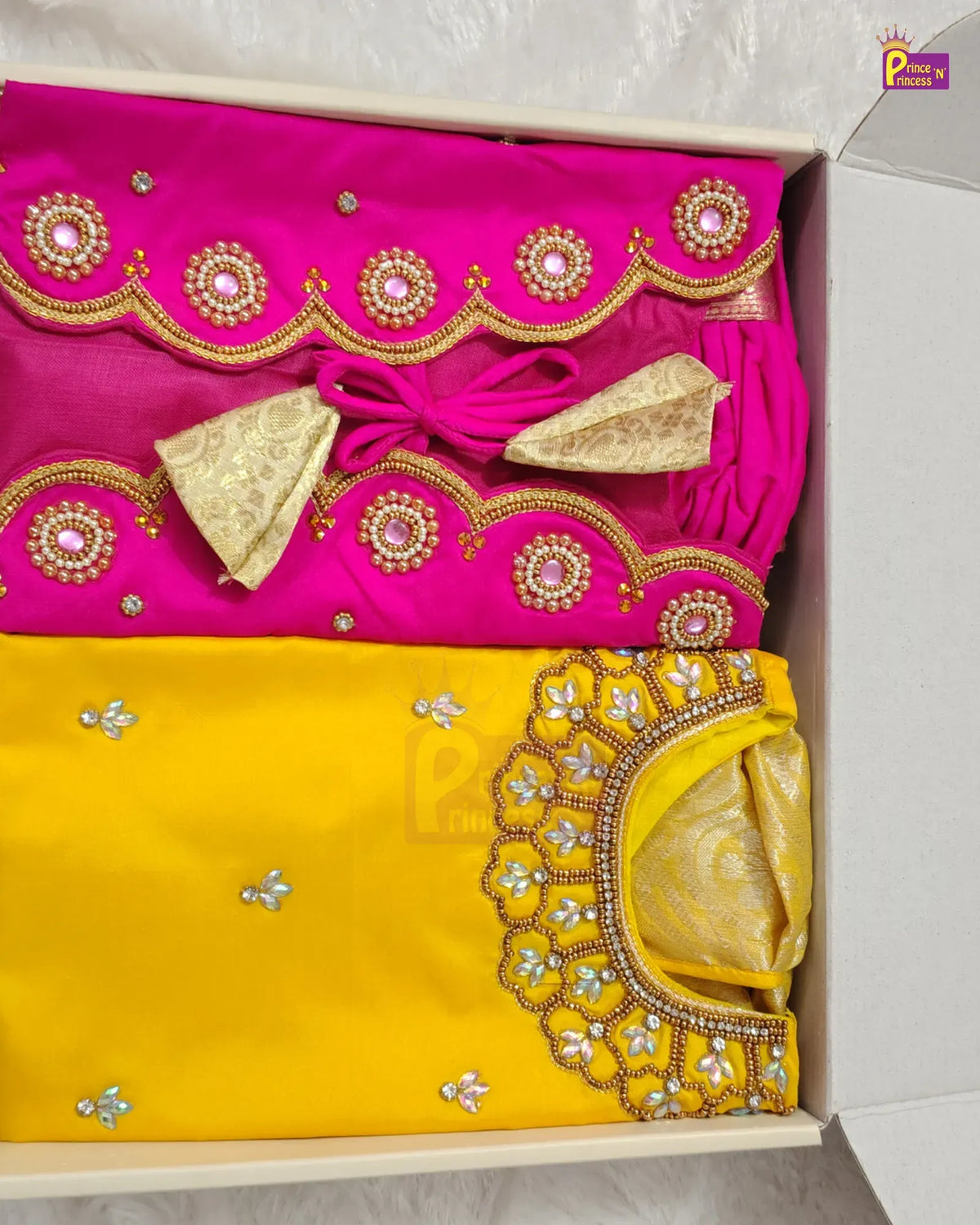 A folded traditional Indian outfit displayed inside a light-colored box, featuring a bright pink top with gold and silver mirror work embroidery and a matching yellow bottom with a heavily embellished scalloped neckline.