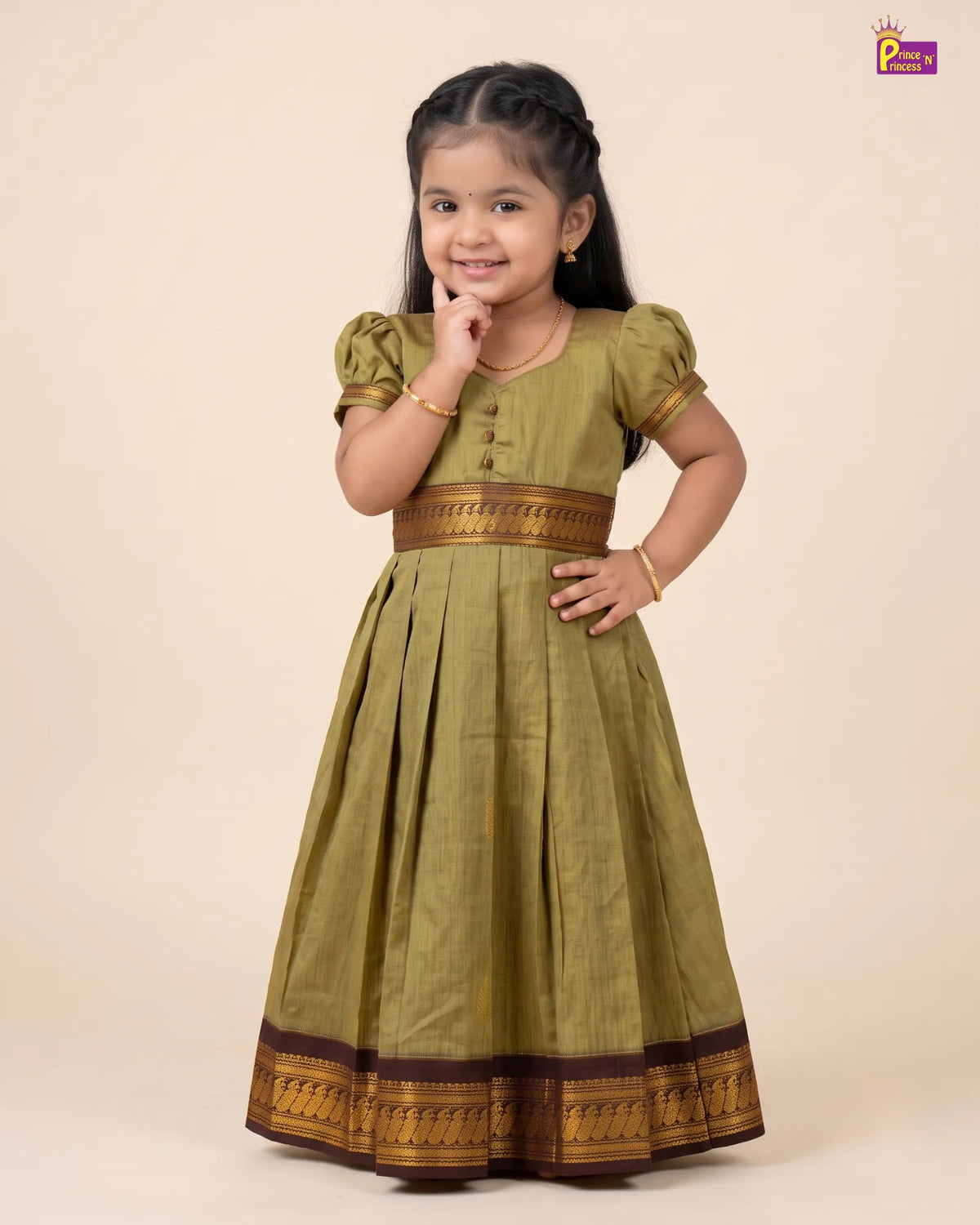 A young girl modeling a traditional Indian dress in olive green with gold and brown borders, standing against a solid beige background.