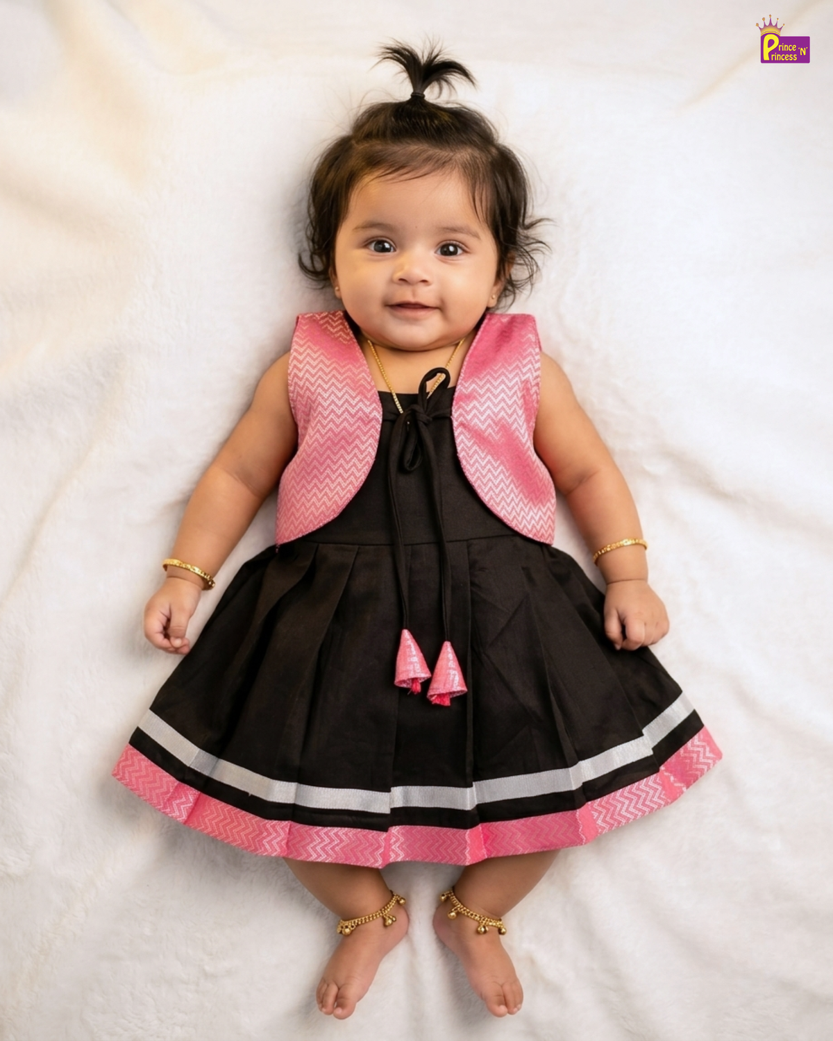 A baby girl wearing a sleeveless black and pink dress with gold jewelry, lying on a white textured blanket.