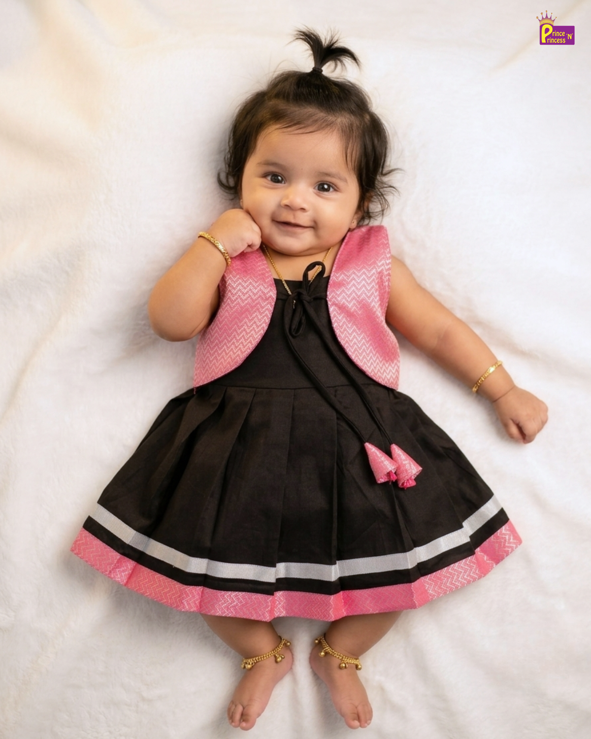 A baby girl wearing a black and pink sleeveless dress with a matching pink vest, lying on a white textured blanket.
