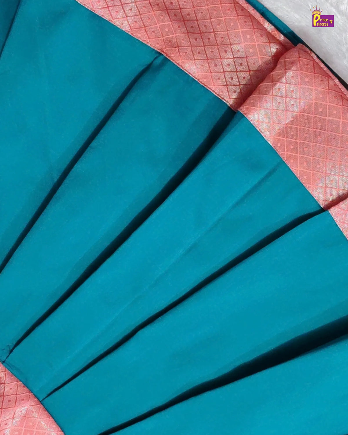 A close-up of a teal blue pleated fabric, likely a sari, featuring a pink border with a subtle geometric pattern.