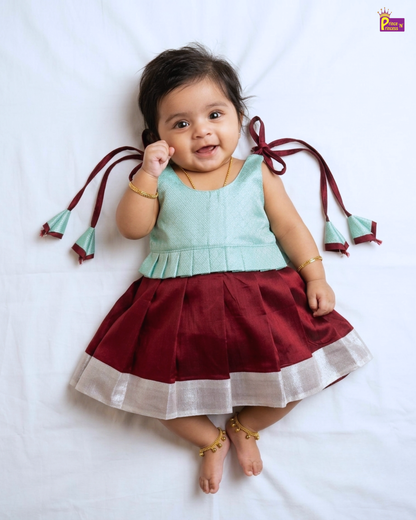 A baby girl lying on a white fabric background, wearing a sleeveless light mint green top and a pleated maroon skirt with a white border and matching maroon ribbon ties on her shoulders.